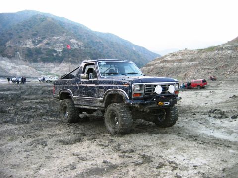 1985 Ford Bronco - A heavily modified Ford Bronco navigates a muddy, off-road terrain with hills in the background.
