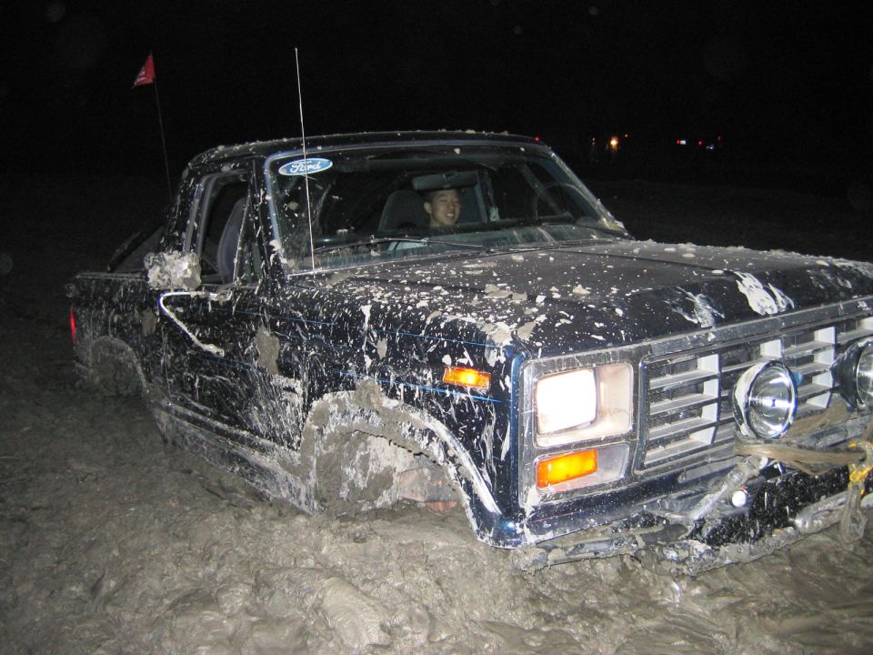 Mud-covered dark Ford truck driving through deep mud at night with driver visible