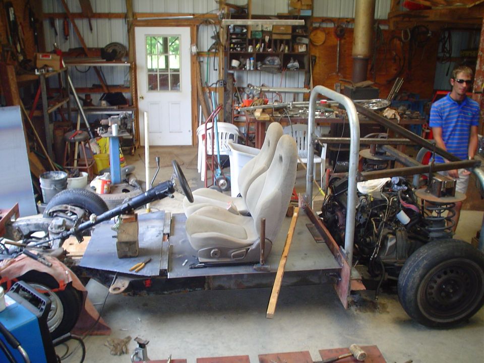 Vehicle chassis with two bucket seats, steering column, and engine in a workshop