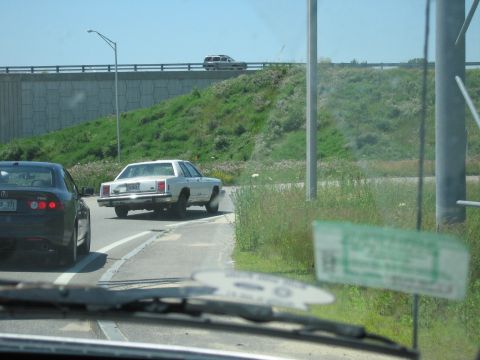 1991 Ford LTD Crown Victoria - Windshield-view photo of a white boxy Ford sedan pulled onto the shoulder of a highway ramp with its rear wheel visibly canted and the body…