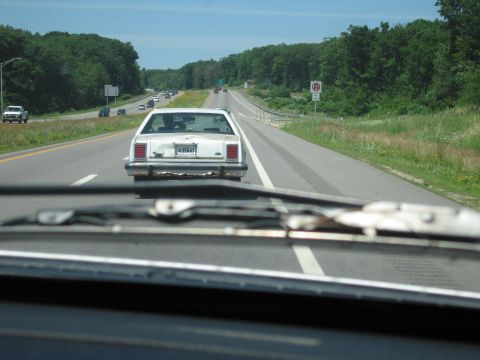 1991 Ford LTD Crown Victoria - Through-the-windshield shot from a following car of a weathered white boxy Ford sedan running down a divided highway, its trunk lid heavily…