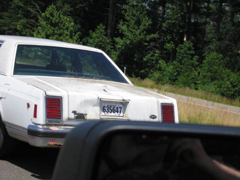 1991 Ford LTD Crown Victoria - Shot from a passing car's window, a weathered white boxy Ford sedan with faded, chalky trunk paint and rust bubbling at the quarter panel t…