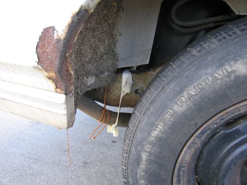 1991 Ford LTD Crown Victoria - Close-up of the rear wheel well on a white car, showing heavy rust perforation at the quarter-panel lip and rocker end, dirt-packed inner f…