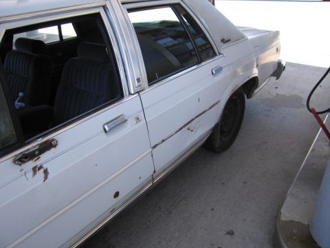 1991 Ford LTD Crown Victoria - Driver-side view of a white four-door sedan parked at a gas pump, showing rust spots, scrapes, and a dent along the doors and rocker panel.…
