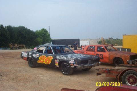 1979 Ford F-150 - Two stock cars are parked on a dirt lot, with trees and construction equipment in the background.