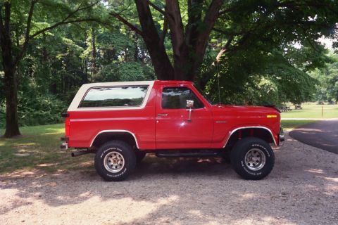 1986 Ford Bronco - This red Ford Bronco is parked on a gravel surface next to a paved road, with trees in the background.