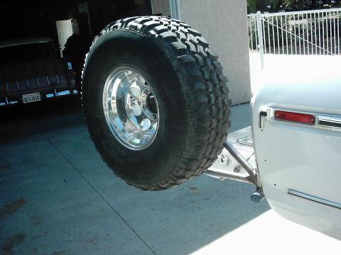 1978 Ford Bronco - A large off-road tire is mounted to a custom rear carrier on a white vehicle, with another vehicle parked in the garage behind it.
