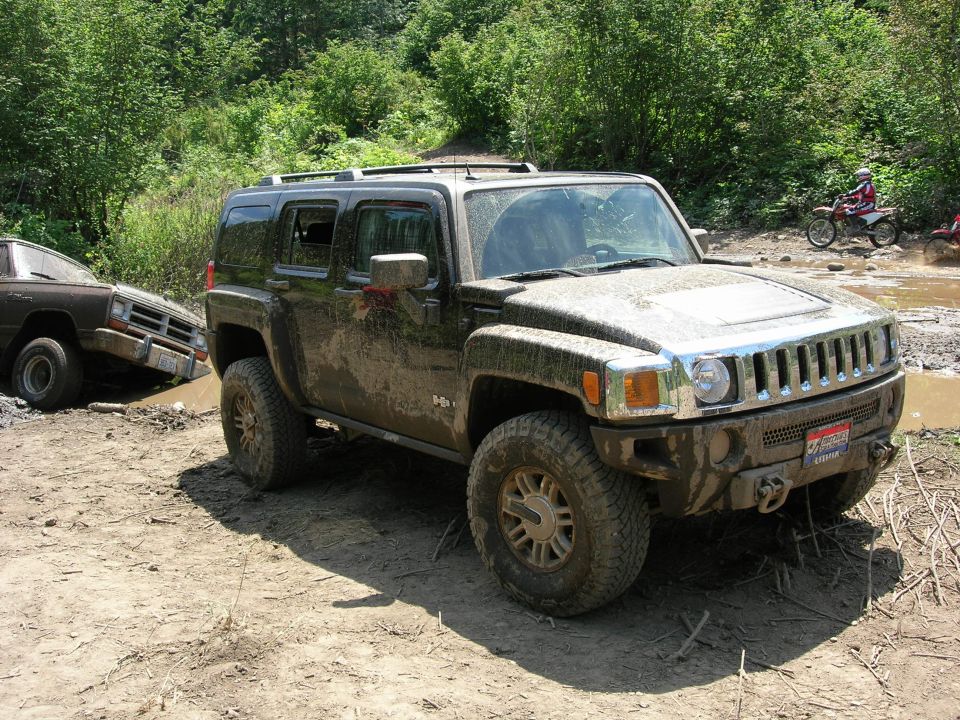 Mud-covered Hummer H3 on a dirt trail with a pickup stuck in mud behind it and dirt bikers in background