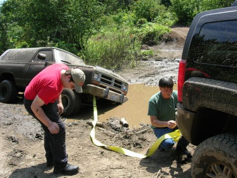 1989 Dodge Power Ram - Two off-road vehicles are stuck in a muddy ditch, with people attempting to use a tow strap to free them.