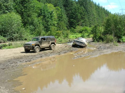 1989 Dodge Power Ram - Two off-road vehicles are stuck in a muddy path surrounded by trees, with one truck partially submerged in water.
