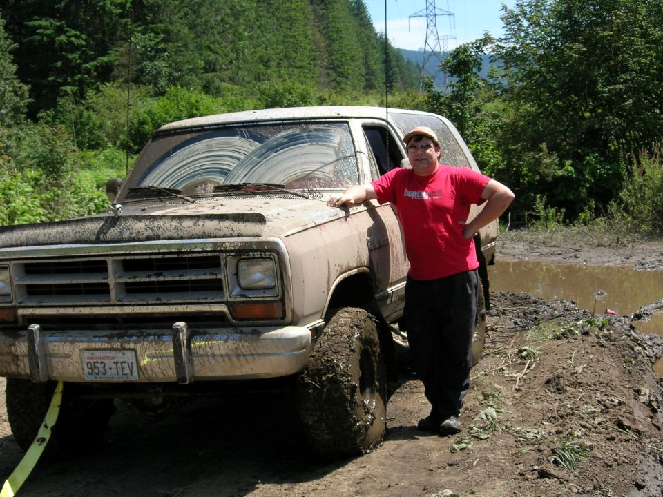 Muddy tan Dodge SUV at 3/4 front angle with man leaning on fender in a muddy off-road setting