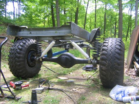 1978 Ford Bronco - Front view of a bare vehicle frame and chassis sitting on large off-road tires in a wooded yard, showing a solid front axle, coil springs,…