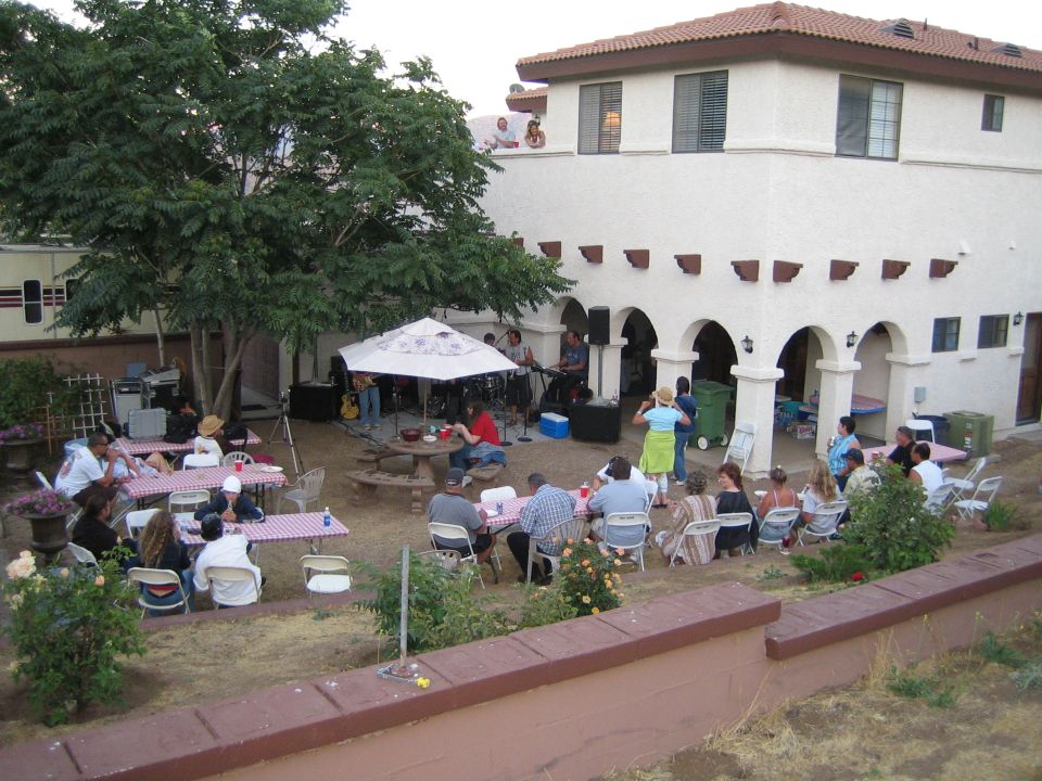 Outdoor party scene with people seated at picnic tables under a tree, a band performing under an umbrella, and a house in the background