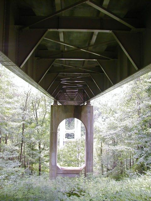 1994 Ford Probe - The intricate metal framework and concrete supports of a bridge are visible from below, with lush greenery on either side.