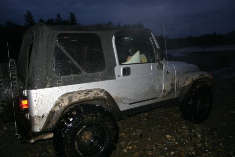 2003 Jeep TJ - A muddy silver 2003 Jeep TJ with a black soft top is parked off-road at dusk.