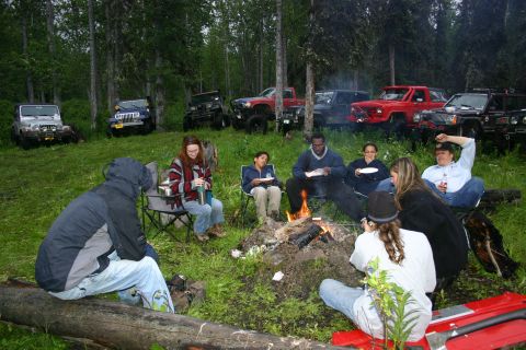 2003 Jeep TJ - A group enjoys a campfire in a forest clearing, surrounded by parked off-road vehicles.
