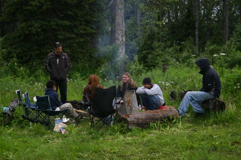 2003 Jeep TJ - A group of individuals gathers around a campfire, enjoying the outdoors amidst lush greenery and tall trees.
