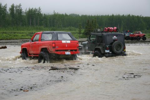 2003 Jeep TJ - Multiple off-road vehicles navigate a muddy riverbed under an overcast sky, with a forest lining the bank.