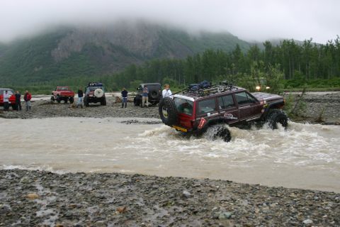 2003 Jeep TJ - Several off-road vehicles navigate a muddy river under a cloudy sky.