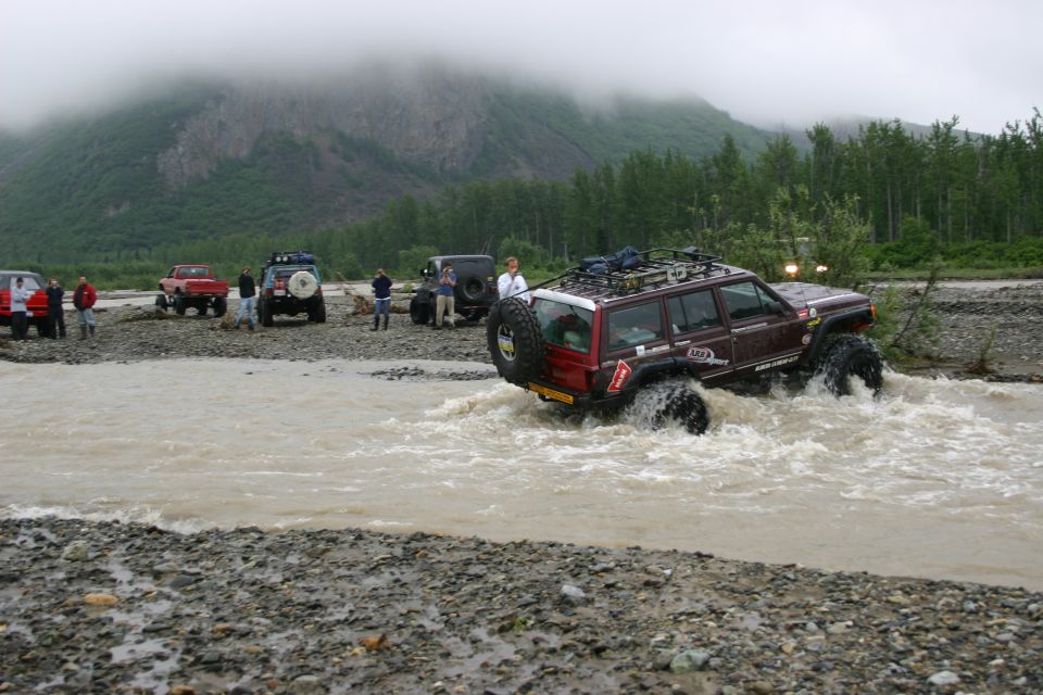Maroon Jeep Cherokee XJ driving through a river, with a spare tire on the back and a roof rack