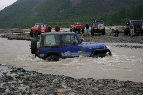 2003 Jeep TJ - A blue 2003 Jeep TJ navigates a rocky riverbed, with several other off-road vehicles and people visible on the shore.