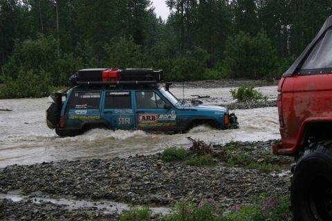 2003 Jeep TJ - Blue Jeep Cherokee XJ navigates a river crossing, with a red vehicle partially visible in the foreground.