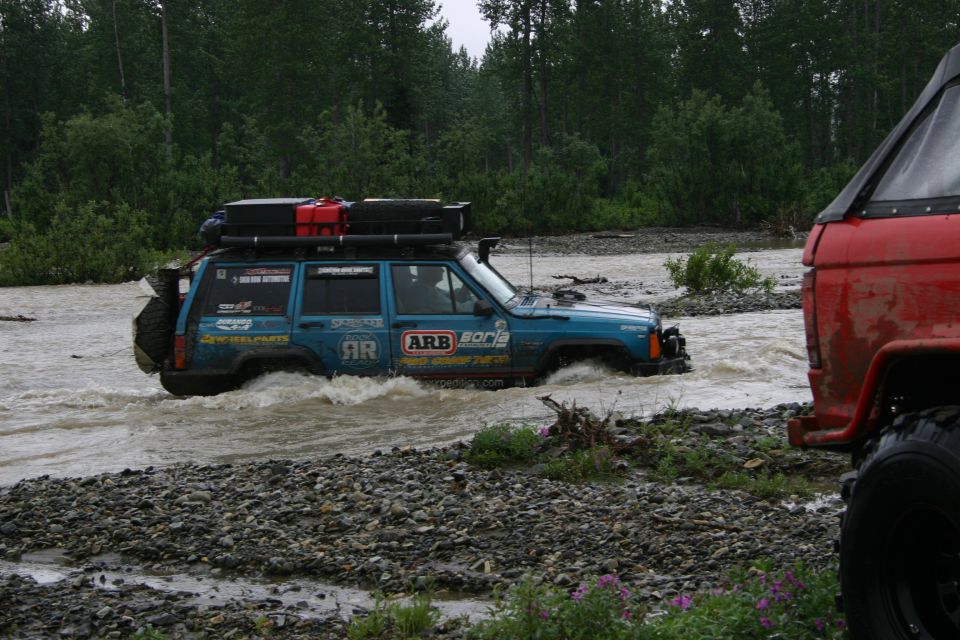 Blue Jeep Cherokee XJ with roof rack and jerry cans driving through river, red vehicle in foreground