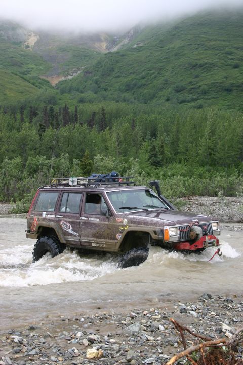 2003 Jeep TJ - A modified Jeep Cherokee XJ navigates a rocky riverbed, surrounded by lush green forest and misty mountains.