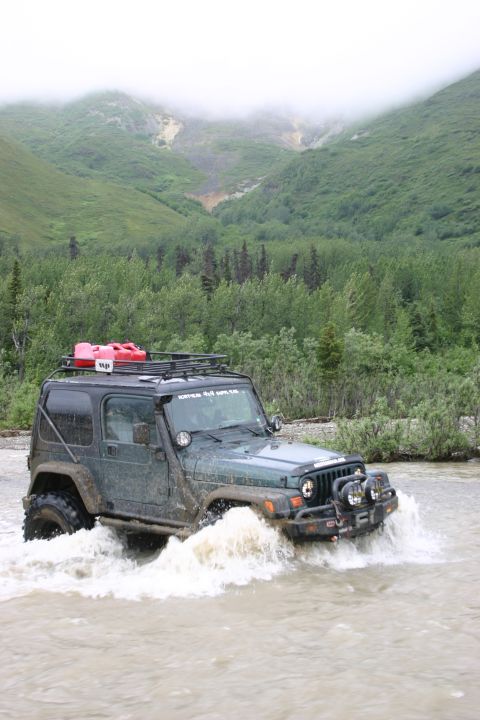 2003 Jeep TJ - A dark green Jeep TJ navigates a shallow river with a mountainous, forested background.