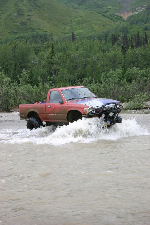 2003 Jeep TJ - A red pickup truck navigates through a river with large off-road tires, kicking up water.