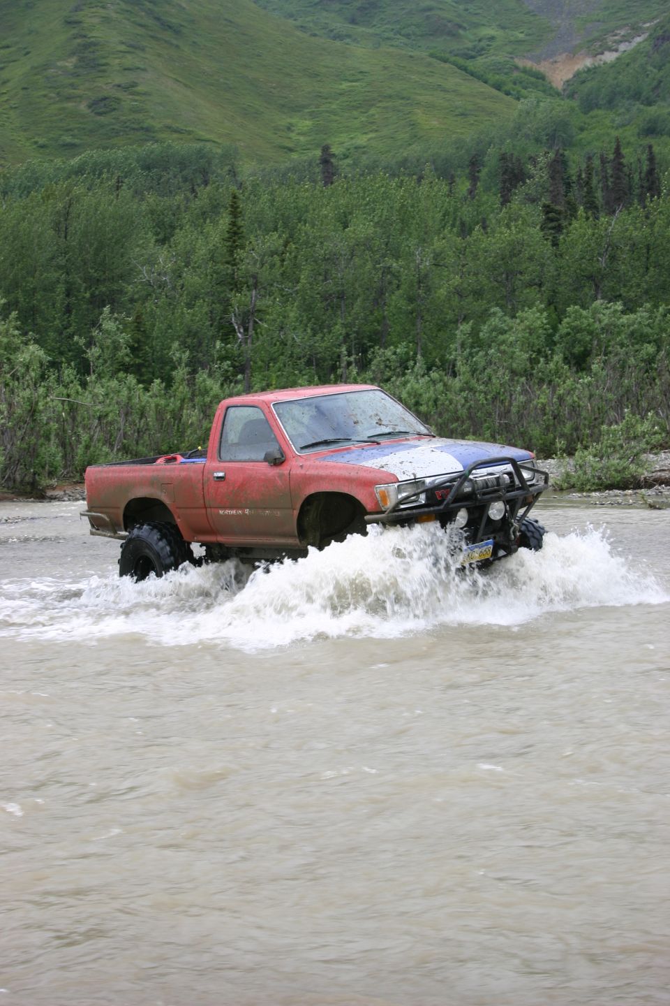 Red pickup truck with off-road tires driving through a shallow river