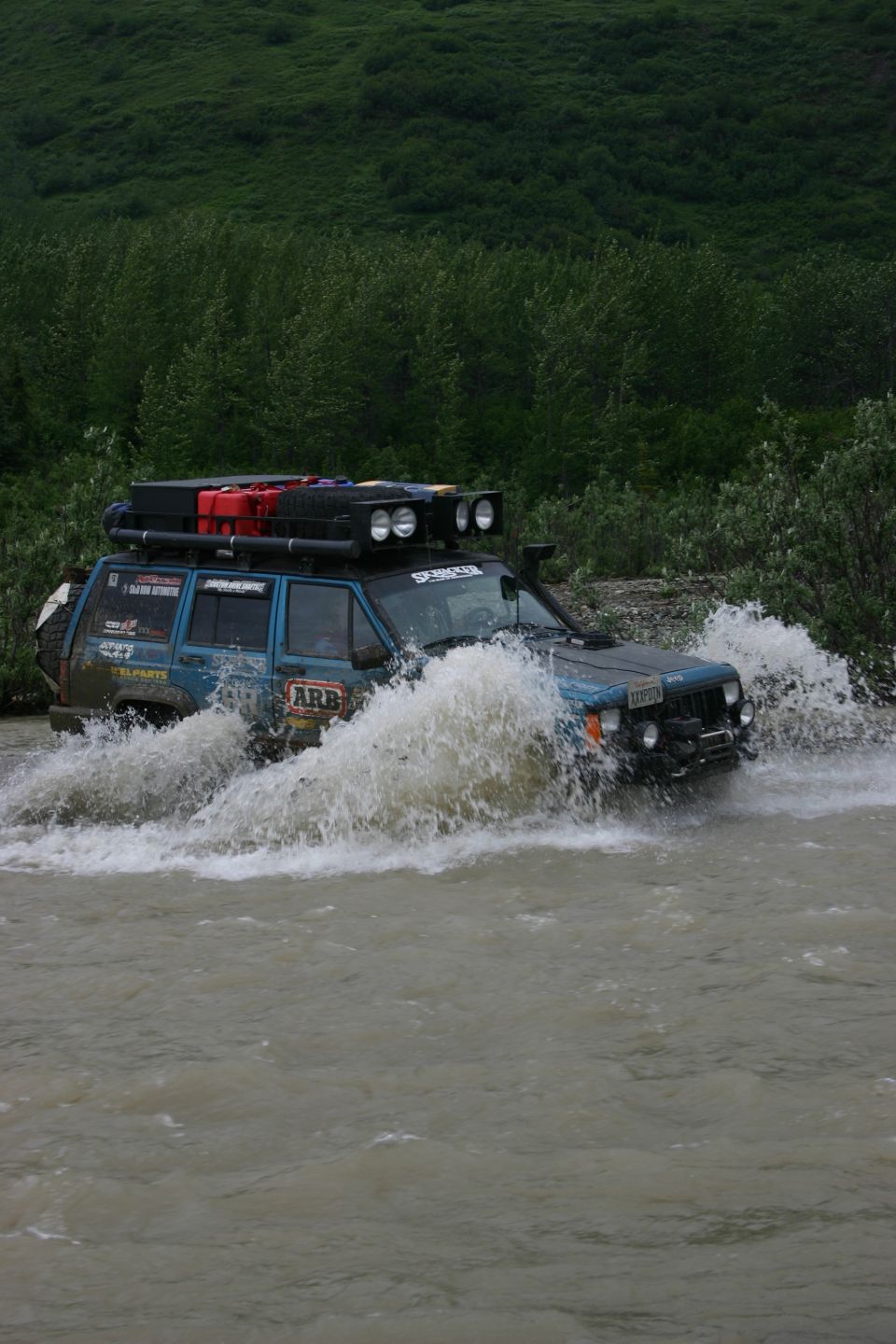 blue Jeep Cherokee XJ driving through water, roof rack with gas cans and lights