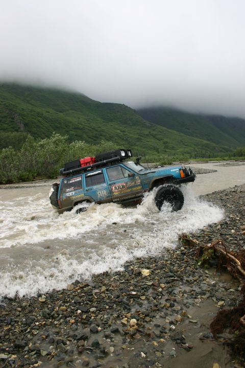 2003 Jeep TJ - A modified blue Jeep Cherokee XJ navigates a rocky riverbed under a cloudy sky.