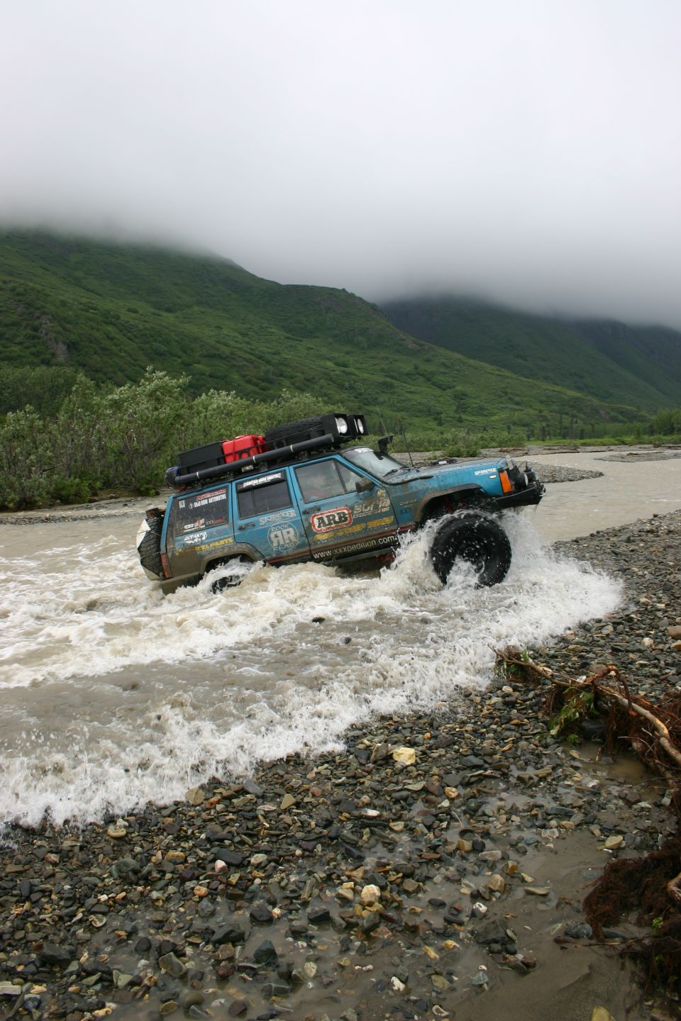 Blue Jeep Cherokee XJ driving through a shallow river, equipped with a roof rack and off-road tires