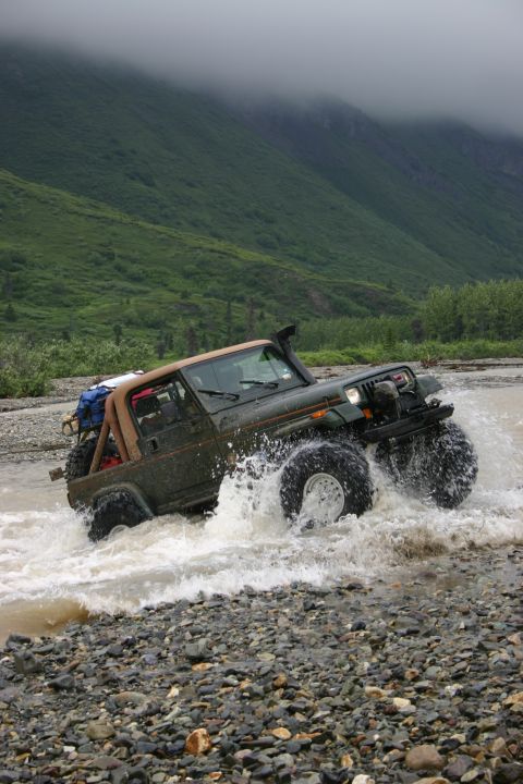 2003 Jeep TJ - A green 2003 Jeep TJ navigates a rocky riverbed, kicking up water.