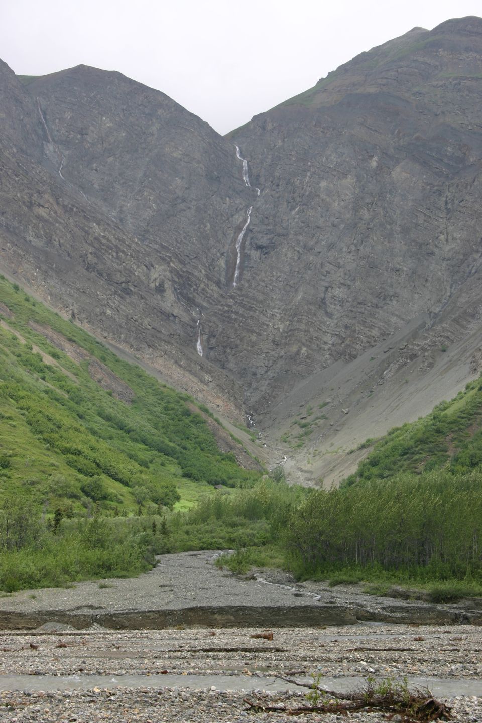 Mountain valley with two waterfalls cascading down a rocky slope, green vegetation on the sides, and a gravel riverbed in the foreground