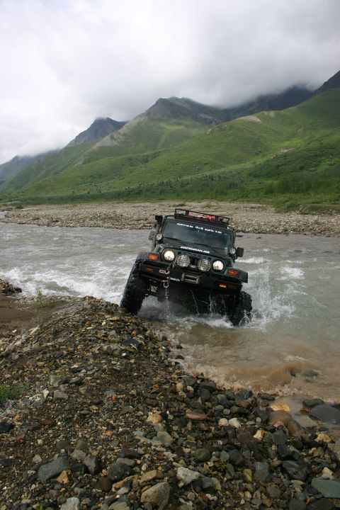 2003 Jeep TJ - A Jeep Wrangler navigates a rocky riverbed under a cloudy sky with mountains in the distance.