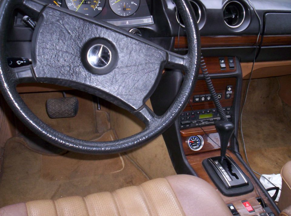 Mercedes-Benz interior with black steering wheel, wood console, automatic shifter, and tan carpet and seats