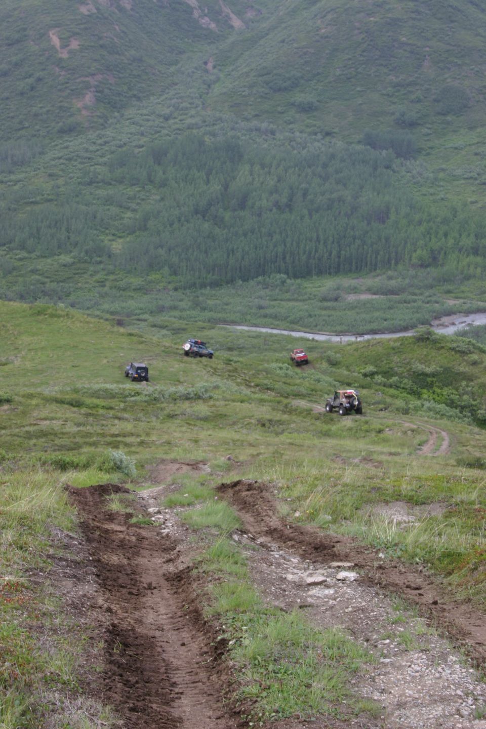 Four off-road vehicles drive on a grassy, uneven terrain with tire tracks visible in the foreground