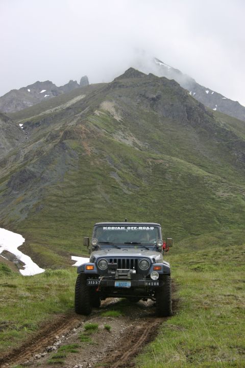 2003 Jeep TJ - A dark blue Jeep Wrangler drives on a muddy track through a grassy landscape with mountains under a cloudy sky.