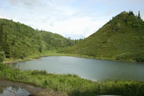2003 Jeep TJ - A calm lake reflects the overcast sky, bordered by lush green hills and vegetation.