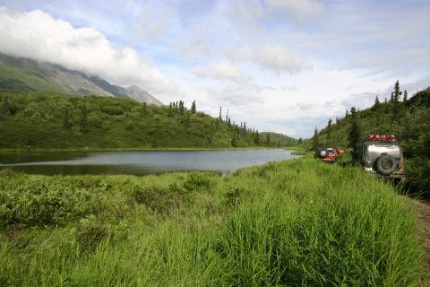 2003 Jeep TJ - Off-road vehicles parked along a lake in a mountainous, wooded area under a cloudy sky.