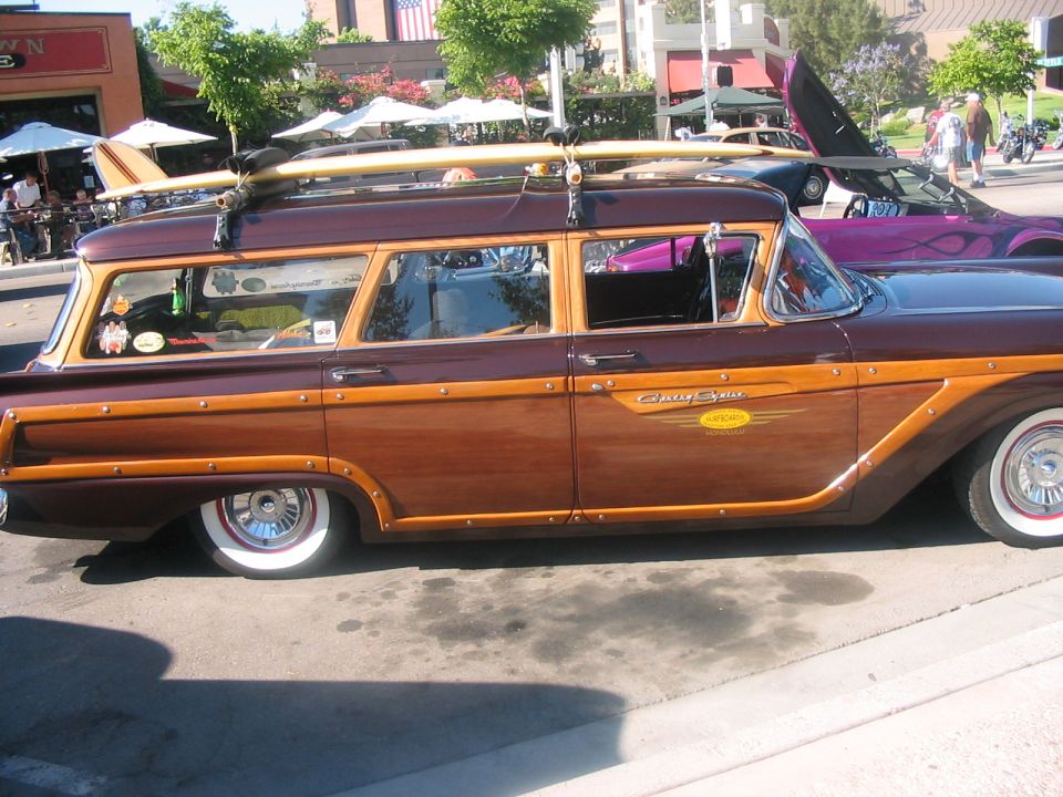 Side view of a 1959 Ford Country Squire station wagon with wood paneling and surfboards on the roof rack. A purple convertible is parked behind it
