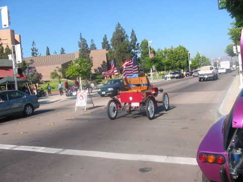1986 Ford Bronco - An antique red car decorated with American flags drives down a street, participating in a parade or event.