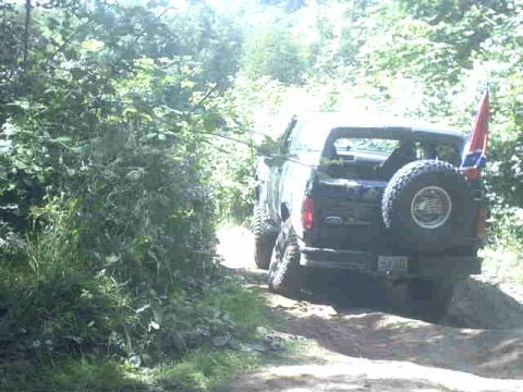 1995 Ford Bronco - A black Ford Bronco drives off-road, displaying a Confederate flag and a spare tire mounted on the back.