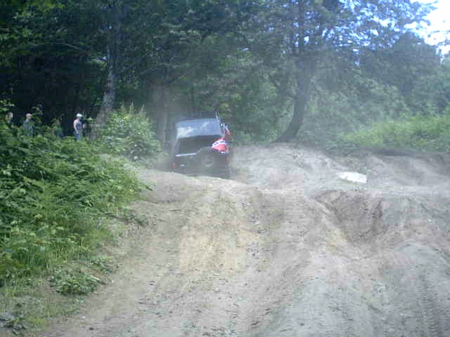 Vehicle kicking up dust climbing a dirt hill on a trail with onlookers