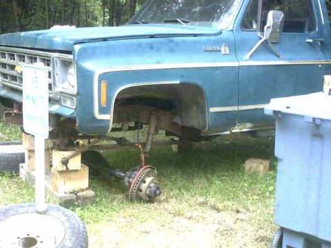 1976 Chevrolet C/K Pickup - A blue pickup truck is shown with its front wheel assembly removed and the vehicle elevated on wooden blocks.