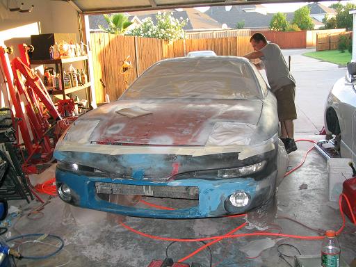 Sedan in garage mid-bodywork with primer, red hoist, and a man working on the roof