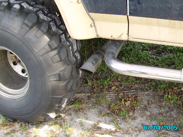 Large off-road tire and chrome side-exit exhaust pipe under a tan Bronco on sandy ground