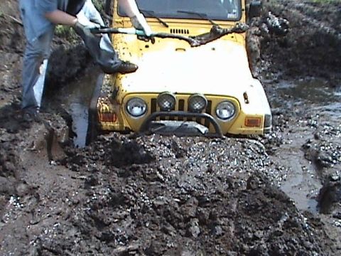 2000 Jeep Wrangler - A yellow Jeep TJ is stuck deep in the mud, with a person attempting to help it get unstuck.